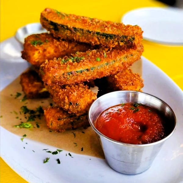 Crispy vegan zucchini fries with herbs and a side of red dipping sauce on a white plate.