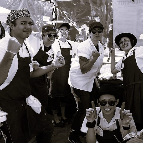 The Wild Chive team, six smiling chefs in aprons and hats, posing with vegan hot dogs.
