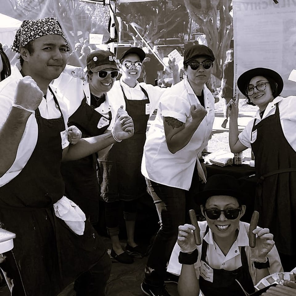 The Wild Chive team, six smiling chefs in aprons and hats, posing with vegan hot dogs.