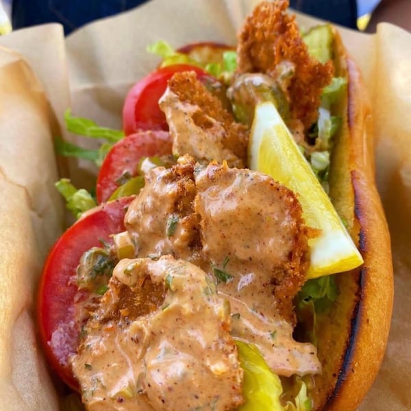 Close-up of a fried seafood po'boy sandwich with lettuce, tomato, pickles, creamy sauce, and a lemon wedge.