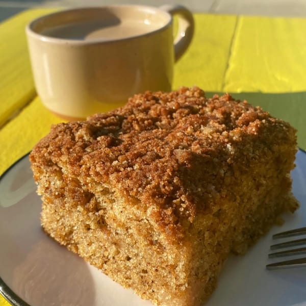 A slice of crumb coffee cake and a coffee mug on a bright yellow outdoor table.